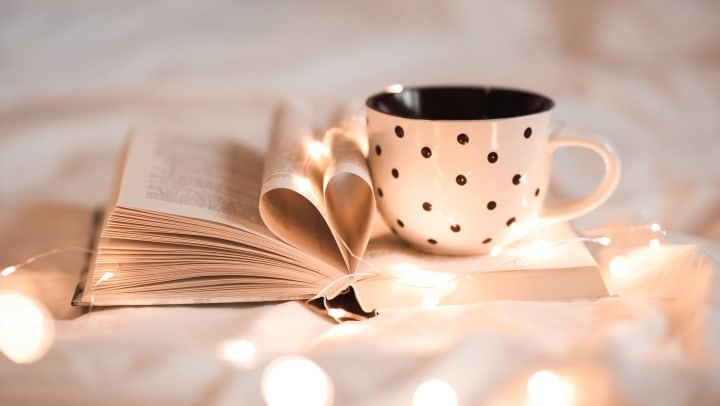 An open book with pages folded into a heart shape with fairy lights and a black-and-white polka dotted mug on top of it. 