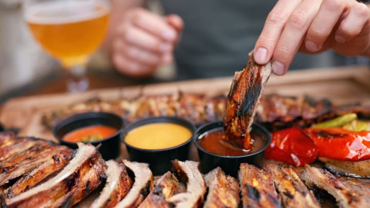 A plate of ribs. A hand is dipping a bbq rib into red sauce. 