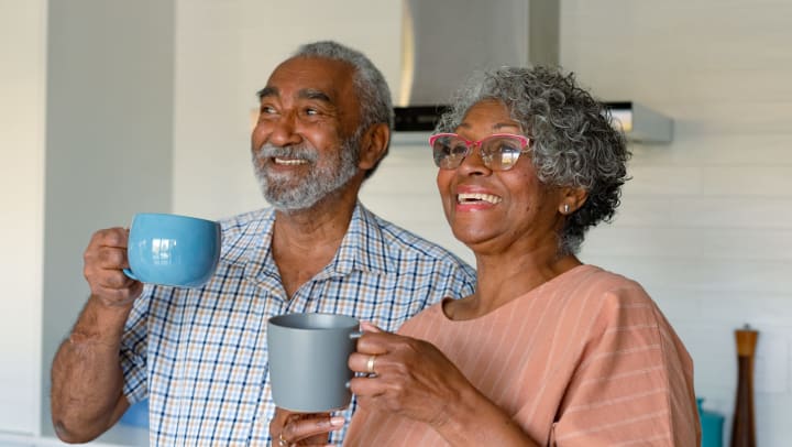 An older couple in a kitchen, smiling and holding coffee cups.