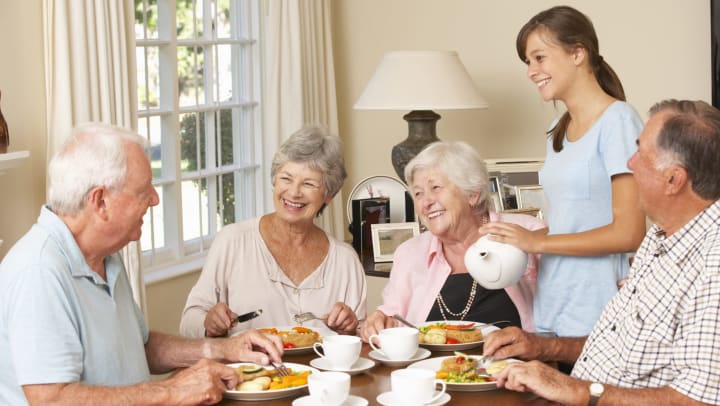 Four seniors sitting at a table in a retirement community with a caregiver serving tea