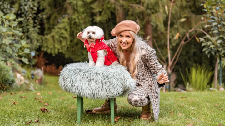 Woman standing behind a little dog in a puffy vest, posing for the camera.