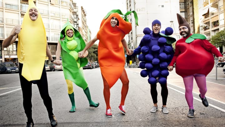 Five men dressed as different fruit and vegetables, holding hands, and smiling on a city street.