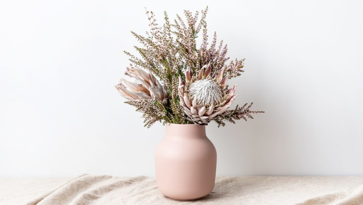 Beautiful floral arrangement including beautiful dried pink King Proteas and delicate thryptomene flowers, in a stylish pink vase, on a rustic table cloth.