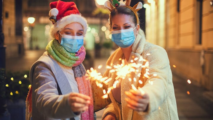 Two young women wearing festive hats and medical masks, hold lit sparklers. 