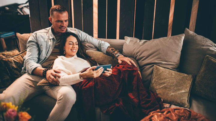 A smiling man and woman lounge on a sofa with velvet pillows and a plush blanket, as they look at her phone. 