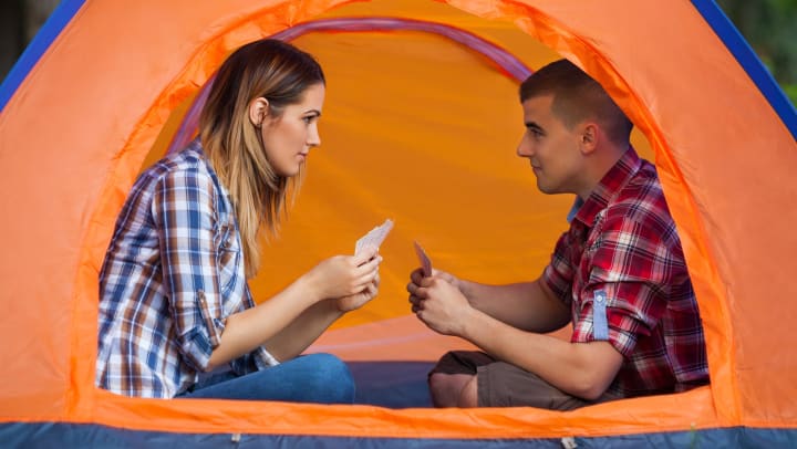 Man and woman in a tent with playing cards in their hands, looking in to each other’s eyes. 