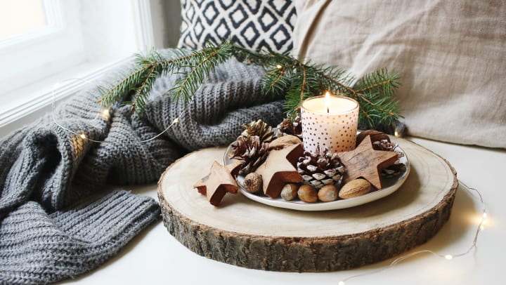 Burning candle decorated by wooden stars, hazelnuts and pine cones placed near window on wooden cut board. Glittering Christmas lights. Fir branch on wool plaid. 