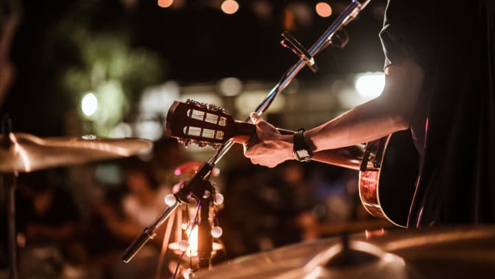 Close up of a musician playing a guitar, with cymbals and a mic stand in the background.