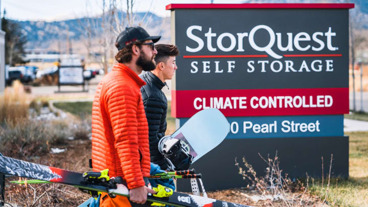 A person carrying skis and a person carrying a snowboard walking in front of a StorQuest Self Storage sign