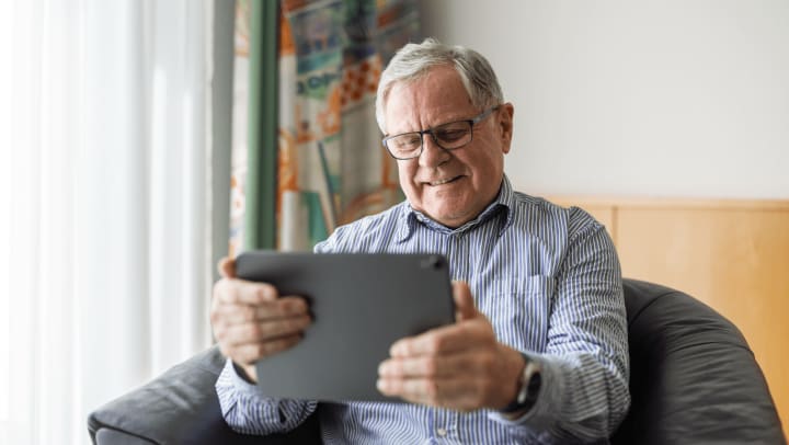 older man in chair holding a tablet and smiling. 
