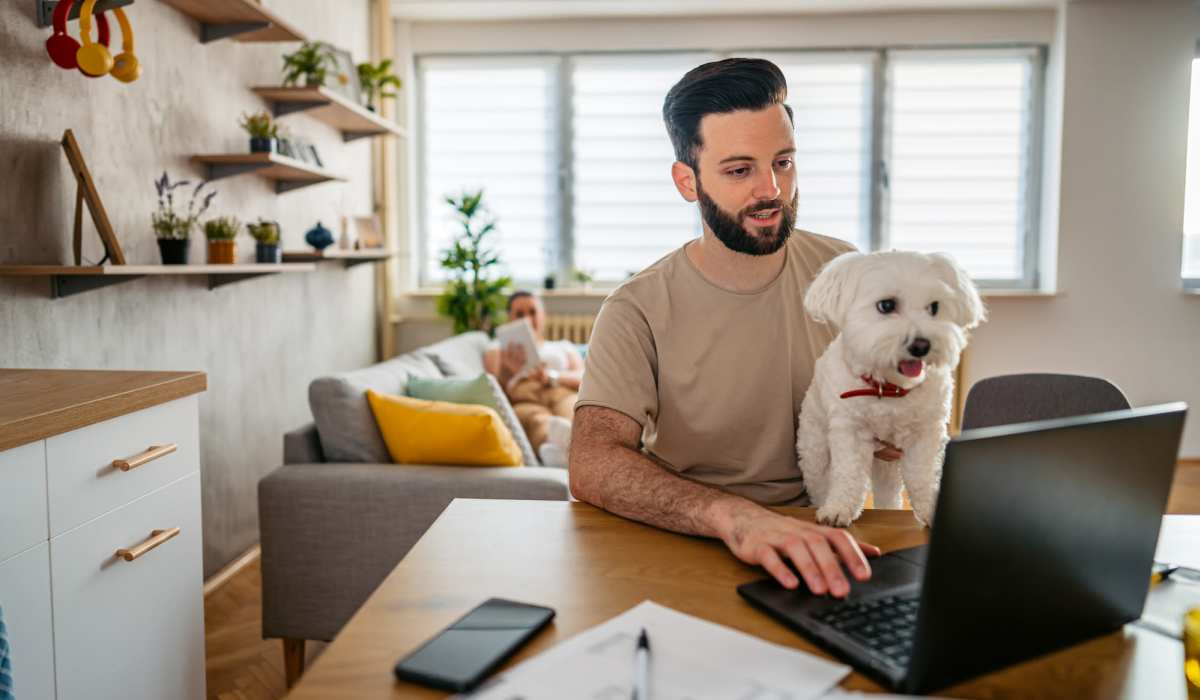 Man working on laptop with small dog in his arms, cozy living room with plants at Dutch Village Apartments in Baltimore, Maryland