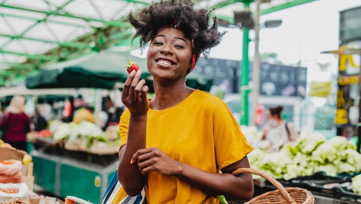 A smiling young woman at a farmers’ market holds up a strawberry in one hand, and holds a shopping basket with some leafy greens in the other.