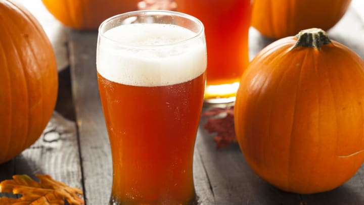 Glass of amber-colored beer sitting on a wooden table surrounded by orange pumpkins and autumn leaves