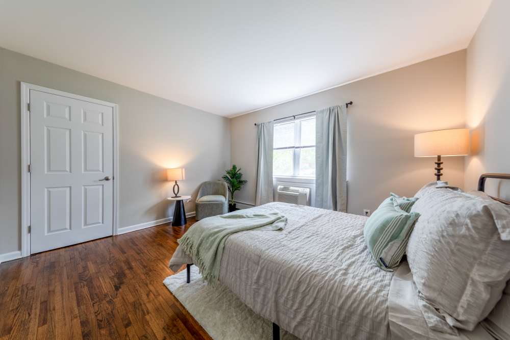 Bedroom with a bed, nightstand with lamp, accent chair, window with curtains, and hardwood flooring at Center Grove Village in Randolph, New Jersey.