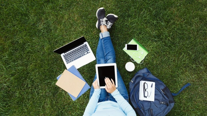 Top view of a young woman in casual outfit using a digital tablet while sitting on grass with laptop, notebook, smartphone, and taking away coffee.