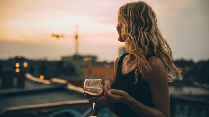 A youthful woman having wine, looking away into the distance while on a rooftop terrace during sunset.