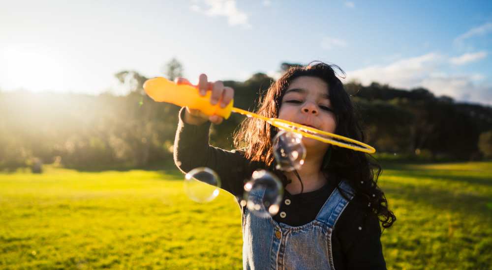 Kid blowing soap bubbles with a bubble wand in a sunlit grassy field near Athena Apartments in Benbrook, Texas