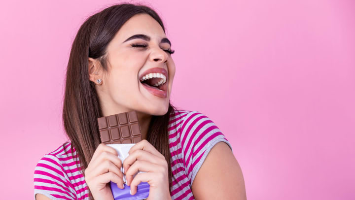 Woman smiling and holding a chocolate bar on a pink background.