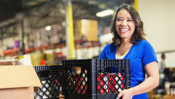 Woman in warehouse smiling and holding a plastic crate full of cans. 