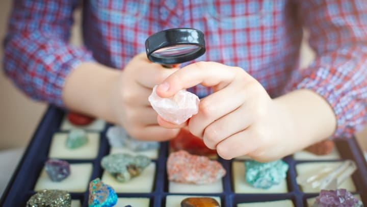 Person in a checkered shirt holding a rose quartz stone and a magnifying glass over a collection of minerals. 