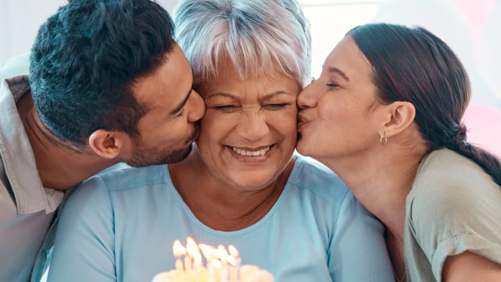 Two adults kissing the cheek of a senior loved one in front of a birthday cake. 