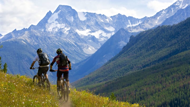 Rear view of two male mountain bikers on a hillside trail with mountains in the background. 