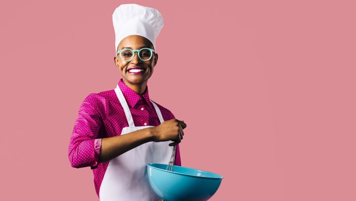 Person in a chef’s hat, pink shirt, and mint-green glasses holding a mixing bowl and whisk and smiling at the camera. 