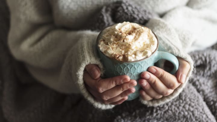 A woman wearing a warm sweater with a fleece blanket over her lap, warms her hands holding a mug of hot chocolate with whipped cream.