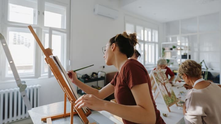 An instructor uses a small paint brush to demonstrate a technique on a canvas in front of a table of students who are all painting.