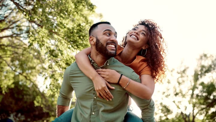 A young man gives young woman a piggyback ride amidst the greenery of a park