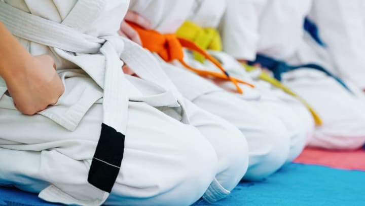 Four people sit in a row at a martial arts class, dressed in white robes and with different colored belts on