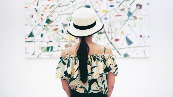 A woman faces away from the camera, looking at a large painting in a gallery