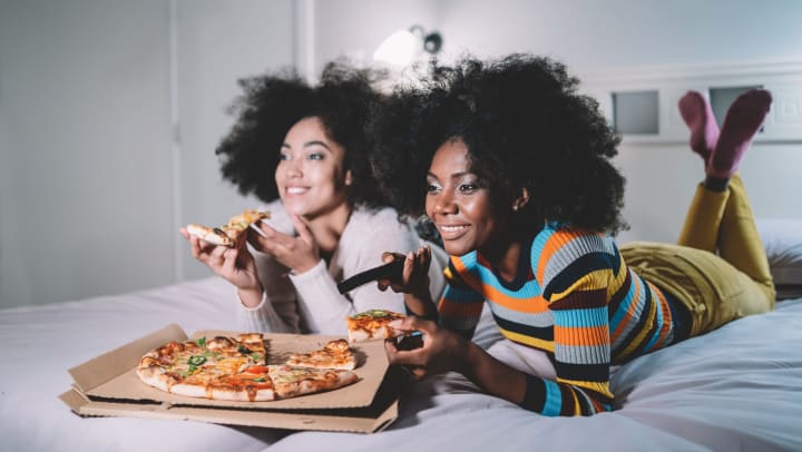 Two women eating pizza while lying on a bed.