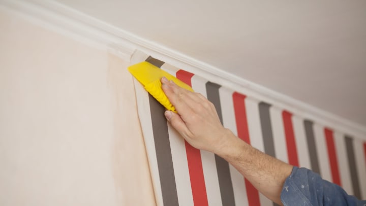 Close-up of a hand smoothing wallpaper with a plastic tool, as it’s applied to a wall.