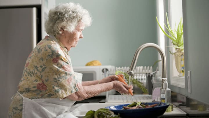 Senior woman with white curly hair cleaning vegetables at a kitchen sink
