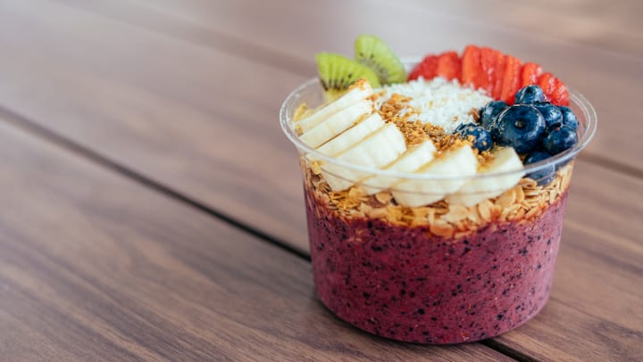 An acai bowl with various fruits in a clear container sitting on a wooden counter. 