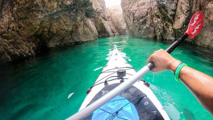 An image of a person paddling a kayak and holding a paddle in a channel with aqua-colored water and rocks in the background.