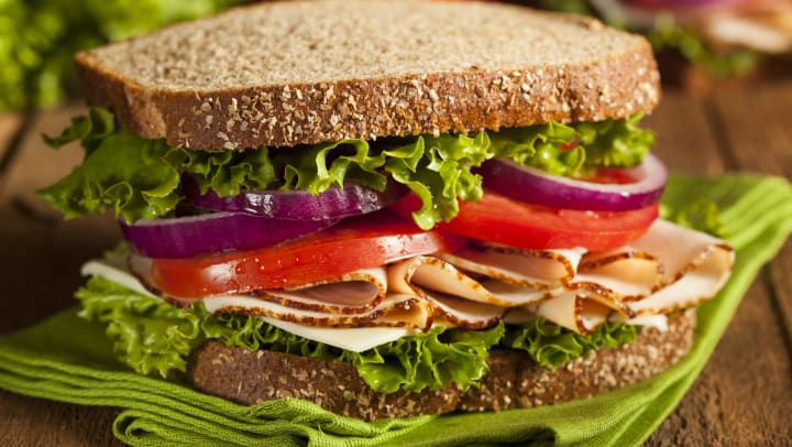 Close up of a sandwich with sliced turkey, lettuce, tomato and red onion sitting on a green plate with vegetables out of focus in the background. 