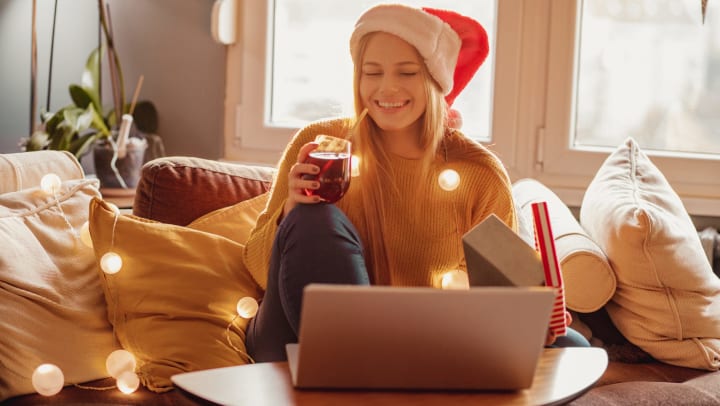 Woman in a Santa hat holding a glass of wine and a gift while smiling into her open laptop computer