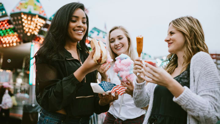 A group of smiling girlfriends enjoying cotton candy, corn dogs and hot dogs at their local county fair.