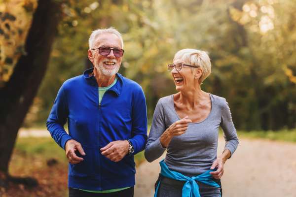 Two seniors jog along a tree-lined path at Watermere Highlands, smiling and enjoying the outdoors in athletic wear.