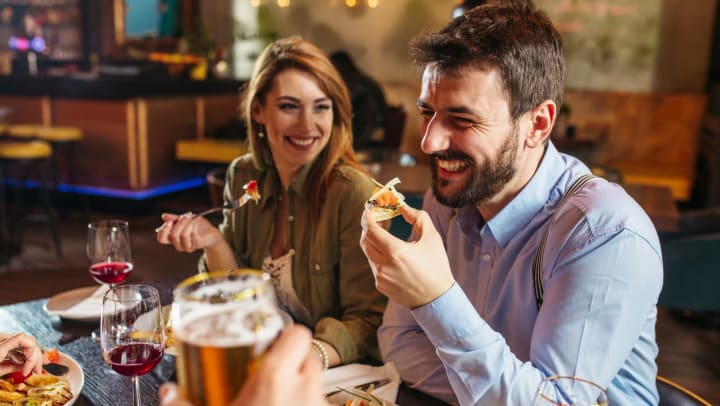 a man in a blue dress shirt laughs as he eats a plate of tapas while a smiling young woman in a green jacket looks on | small bites in Ooltewah