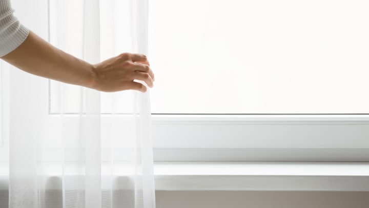 An adult hand is touching sheer curtains in front of a brightly lit window.
