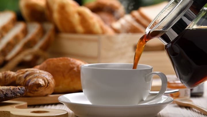 Pouring coffee with steam rising from a cup, with bread/pastries on white wooden table