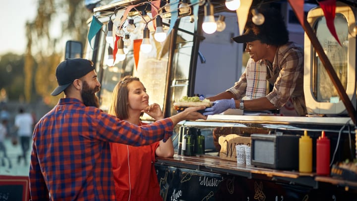 A couple taking sandwiches from a friendly employee through a food truck window.