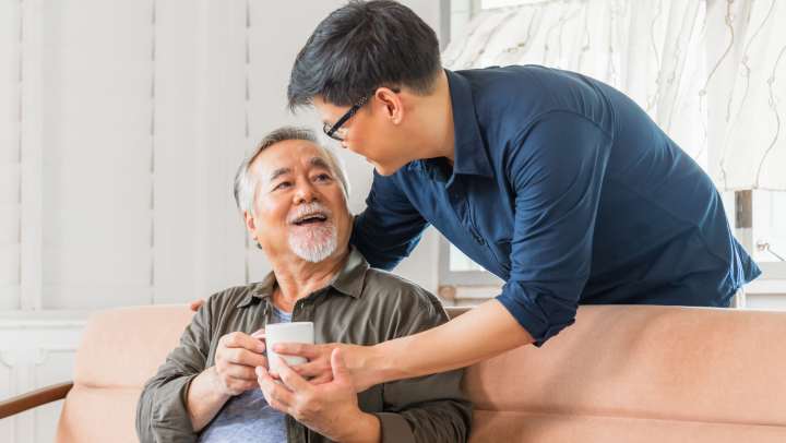 Adult son smiling and giving a cup of drink to his elderly father