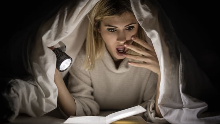 Woman holding a flashlight with a blanket over her head making a shocked face at an open book.