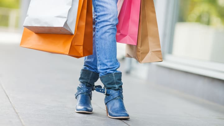 Close up of a woman’s legs in jeans and boots, surrounded by several shopping bags she’s carrying. 