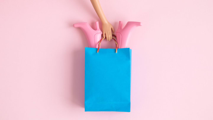 Doll’s arm holding shopping bag with footwear over pastel pink background