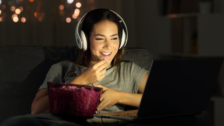 A with headphones on watching something on a laptop while eating popcorn. 
